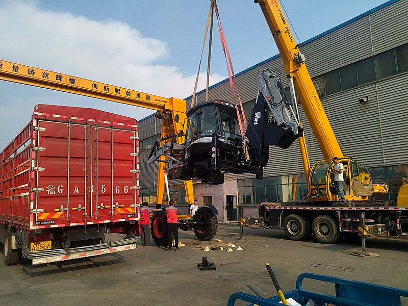 Crane lifting a backhoe loader onto a red container truck under the gantry